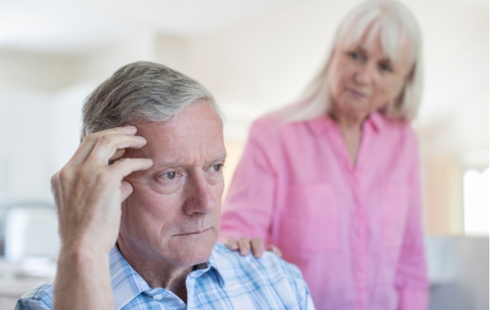 A senior woman looking at her husband while she touches his shoulder with concern as he suffers from Alzheimer’s