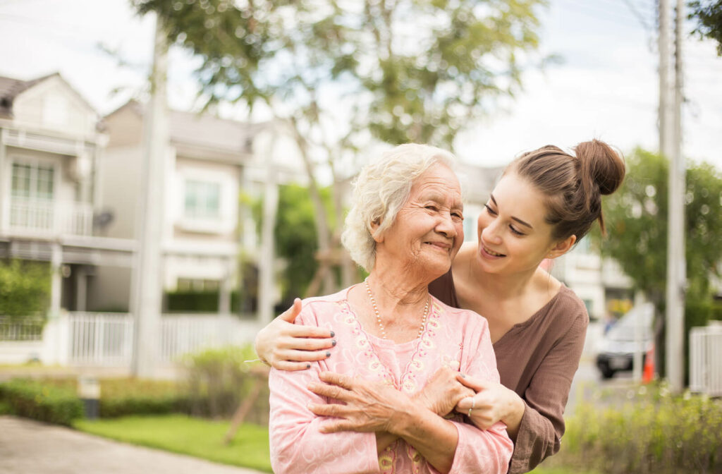 a young woman supports a senior woman from behind with a care home in the background