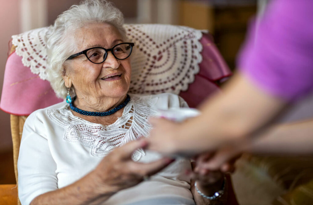 A senior woman with white hair and glasses smiling while holding a caregiver's hand.