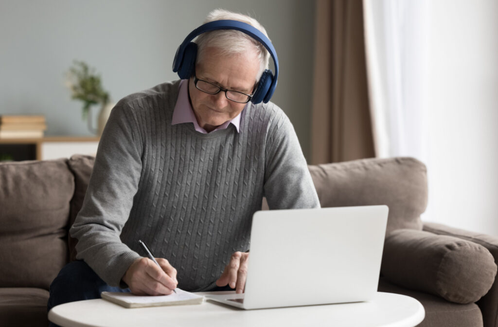 A senior man taking notes in front of his laptop while wearing a wireless headphones.