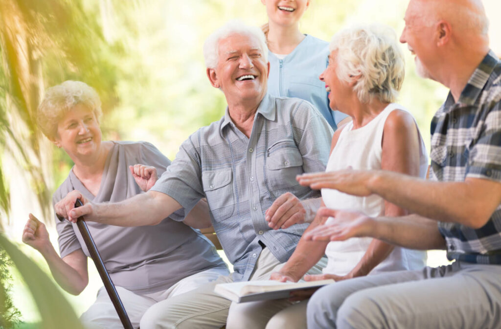An older man with a cane smiles and laughs while sitting outside with a nurse and other older adults